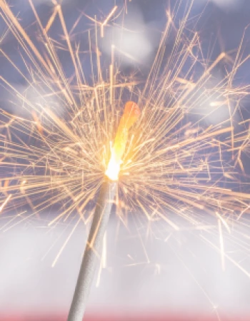 The image shows a lit sparkler emitting bright sparks, with a blurred background that appears to be the American flag.